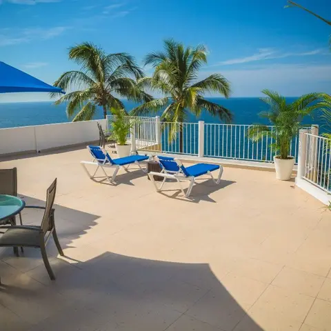 Rooftop terrace with a blue market umbrella and dining set, two blue-cushioned loungers, potted palms, and an open sea view beyond the railing
