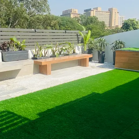 Rooftop terrace with artificial turf, a built-in stone bench and a floating teak bench, potted banana plants lined along a slatted privacy screen.