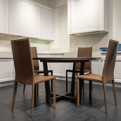 Round dark-wood dining table with four tan leather chairs on dark wood flooring, set against white shaker kitchen cabinetry and a stone slab backsplash.