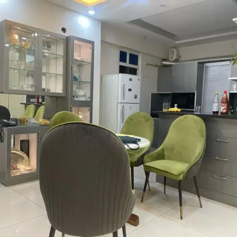 Round dining table with olive-green velvet chairs in front of a glass-front crockery display unit, kitchen visible behind