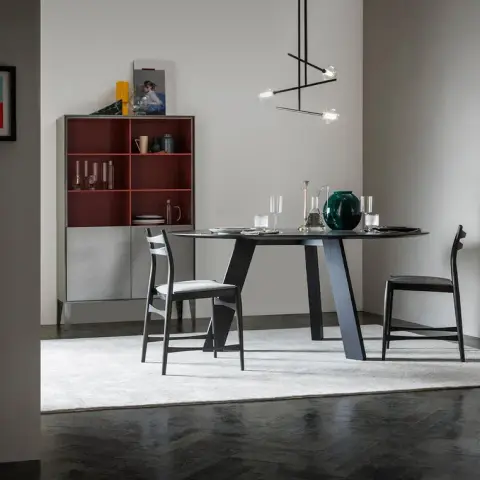 Round ebonised dining table on a faceted trestle base with two ladder-back chairs, set on a pale rug over dark herringbone parquet