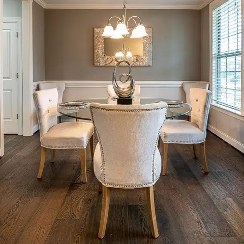Round glass-top dining table with upholstered nailhead chairs beneath a crystal chandelier and a framed mirror on a wainscoted wall.