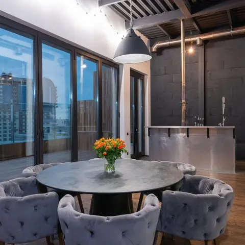 Round marble dining table with tufted velvet chairs under an industrial pendant, exposed ceiling ductwork and a dark block feature wall against