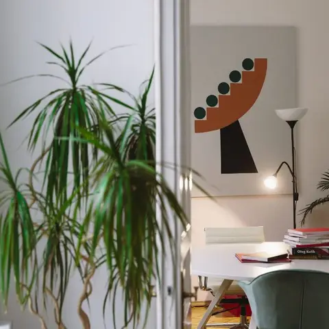 Round white desk with a teal velvet chair and floor lamp, an abstract color-blocked poster on the wall, framed by large potted plants.