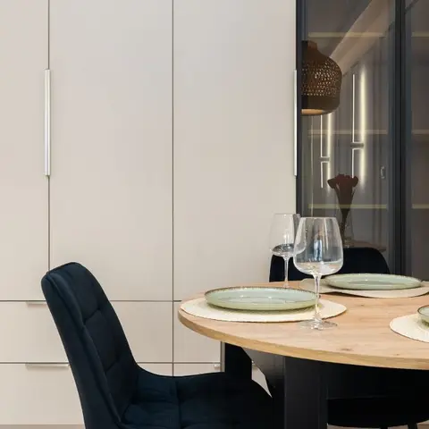 Round wood dining table set with sage-green stoneware and wine glasses, black velvet chairs, and a tall cabinet wall beside a glass-front bar nook.