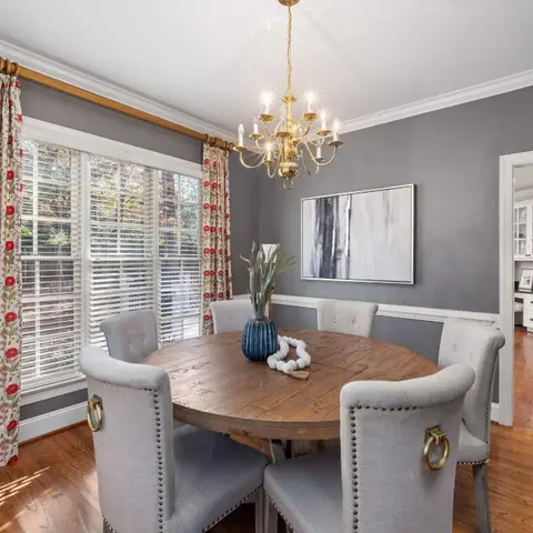 Round wooden dining table with grey nailhead-trim upholstered chairs beneath a brass candelabra chandelier, floral drapes on the window behind