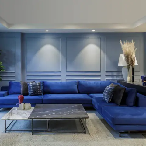 Royal-blue velvet L-sofa running the length of a blue-grey moulded panel wall lit by wall-wash spots