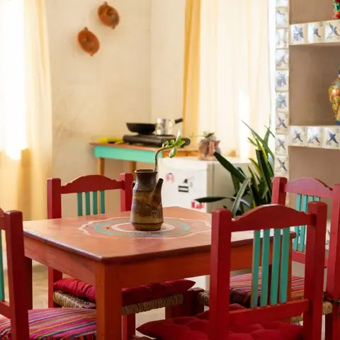 Rustic dining nook with a hand-painted red table, teal-and-red woven-seat chairs, and a blue-and-white patterned tile display niche in the corner.