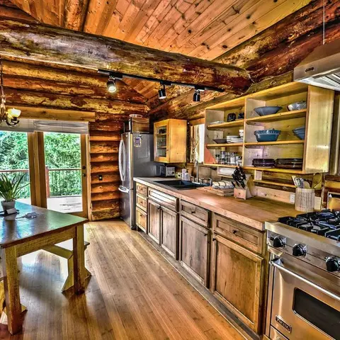 Rustic log-cabin open kitchen and dining space with exposed log walls, open wood shelving and a stainless steel range
