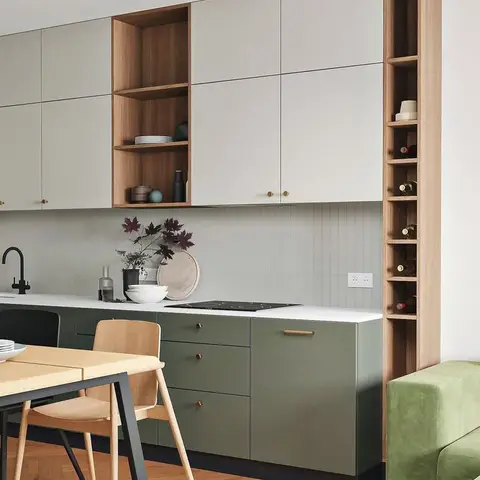 Sage green and off-white two-tone kitchen with an open wood shelving and wine-storage tall unit, wood dining table and chairs in the foreground.