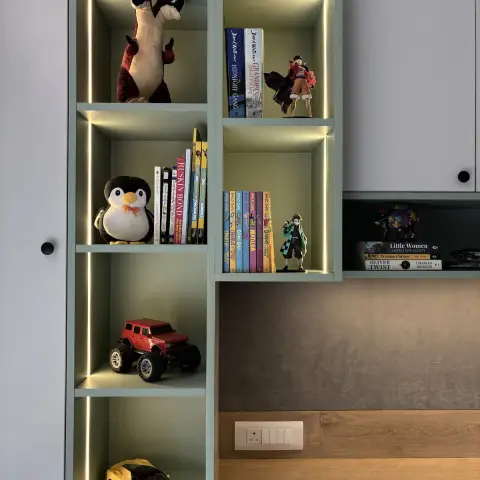 Sage-green backlit display shelving over a study nook, styled with children's books and toy figures against a grey suede-effect wall.