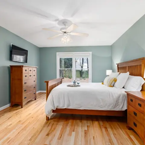 Sage-green bedroom with a mission-style wooden bed and matching tall dresser, wide-plank oak flooring, and a wall-mounted TV opposite the bed.