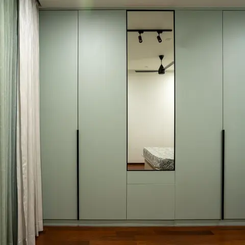 Sage-green floor-to-ceiling wardrobe wall with a full-length mirrored door reflecting the patterned bedspread and ceiling fan of the room beyond.