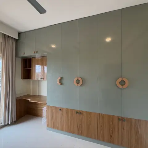 Sage-green glossy wardrobe wall with wood-trim doors and circular pull handles, beside a built-in wood study desk with open shelving by a curtained window.