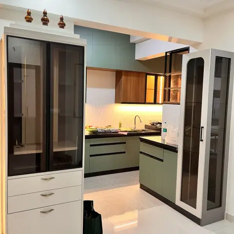 Sage-green kitchen viewed through the gap between two tall bronze-glass crockery towers, with teak-laminate wall units