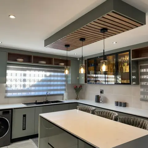 Sage-green kitchen with a large white-topped island seating three on tufted stools, under a dropped wood-slat ceiling feature with three pendant lights.