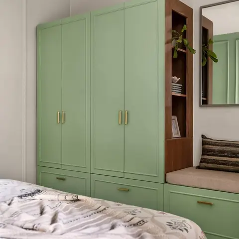 Sage-green shaker-profile wardrobe run with slim brass bar pulls, broken by a walnut open-shelf tower and a cushioned window-seat bench over green drawer