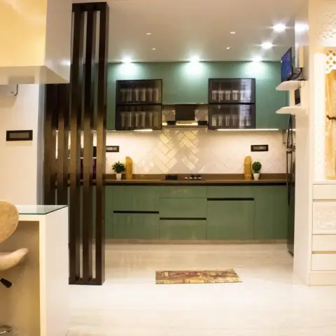Sage-green straight-run modular kitchen with white herringbone dado, framed by a dark fluted timber partition on the left and a white CNC-jaali pooja unit on