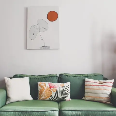 Sage-green tufted sofa styled with mixed-print cushions beneath a minimalist line-drawn botanical art print, next to an arched wooden ladder shelf.