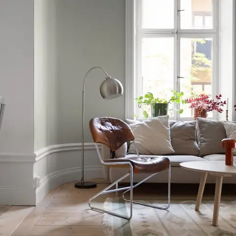 Sage-walled living room corner with a tan leather sling chair beside a curved bay window and a grey linen sofa.