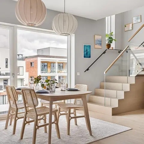 Scandinavian dining nook with a round oak table, woven rattan chairs, and paired paper-lantern pendants beside a light-wood staircase.