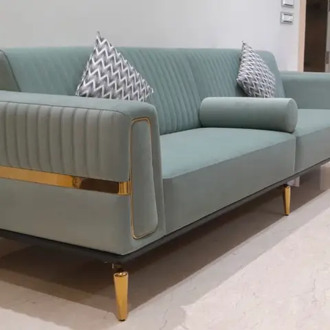 Sea-green channel-tufted three-seater sofa with gold trim and gold conical legs on a marble floor with a hallway visible beyond