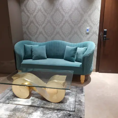 Seating nook with a teal channel-tufted velvet sofa against damask wallpaper, and a sculptural gold infinity-base glass coffee table on a grey rug.