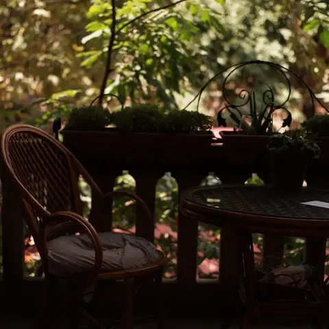 Shaded home balcony with a rattan chair and round table, wrought-iron balustrade and potted plants against a backdrop of dense greenery.
