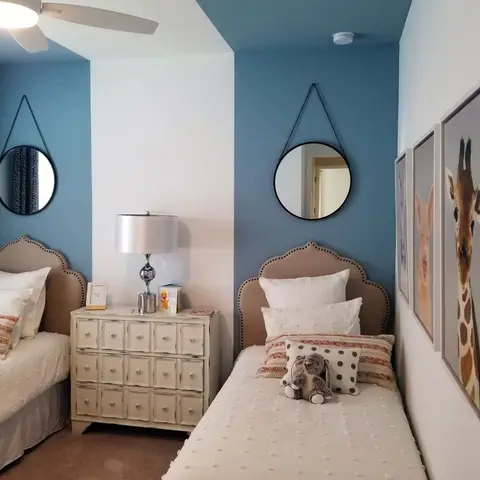 Shared kids' room with two scalloped-headboard twin beds against color-blocked blue and white walls, round hanging mirrors, and a giraffe-and-piglet art trio.