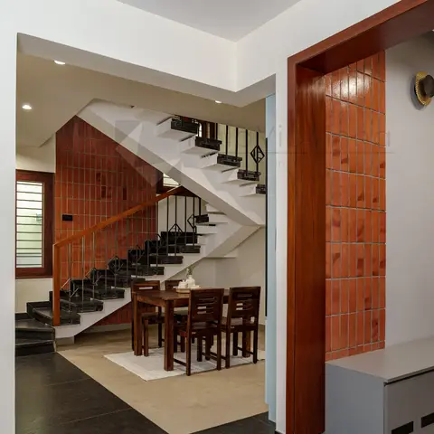 Sheesham dining table for four tucked beneath a dogleg staircase clad in terracotta brick-look tile, dark flooring underfoot.