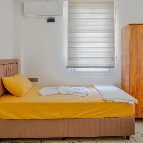 Simple twin-bed guest room with mustard bedding, hotel-style folded towels, a plain wood wardrobe with a mirrored door, and a striped window blind