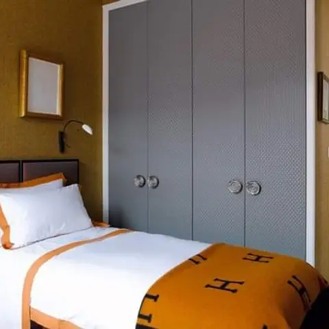 Single bed against mustard textured wallpaper with a gold sunburst mirror, paired with a grey wardrobe with faceted glass knobs and an orange Hermes-print