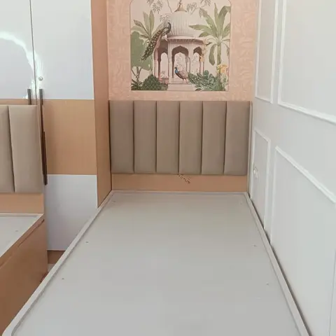 Single storage bed with a fluted taupe upholstered headboard against a pink wallpaper wall featuring a peacock and palace mural, beside a laminate wardrobe.