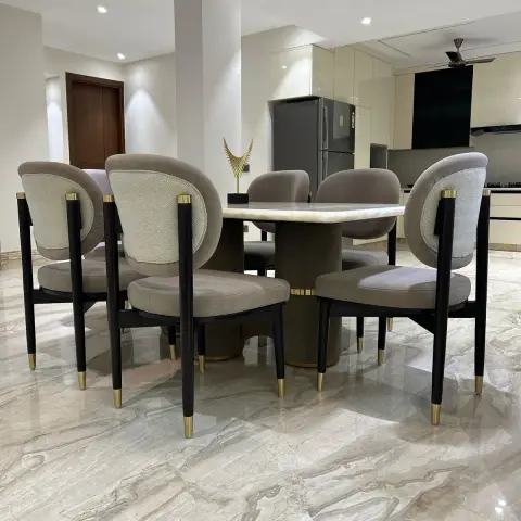 Six round-backed taupe suede chairs on black legs with brass ferrules pulled up to an oval onyx-topped table, with the open kitchen and fridge visible beyond.