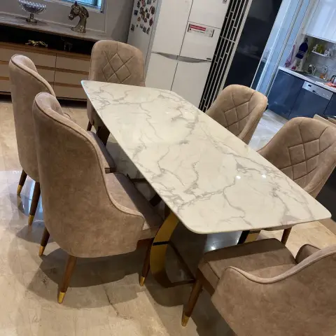 Six-seater dining set on beige marble flooring: a boat-shaped white marble top on a brass X-base, taupe suede chairs with brass-tipped legs