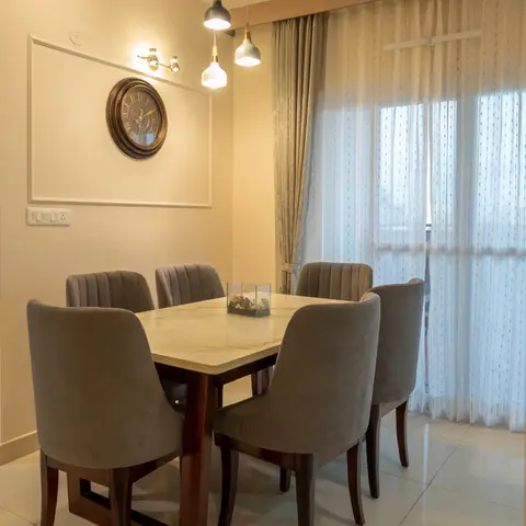 Six-seater dining set with a marble-top table and channel-tufted grey velvet chairs, lit by a cluster of pendant lights beneath a wall clock in a moulded frame.