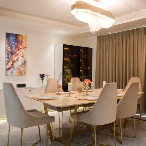 Six-seater marble-top dining table with brass legs beneath a tiered crystal chandelier, facing a fluted glass-and-gold crockery unit.