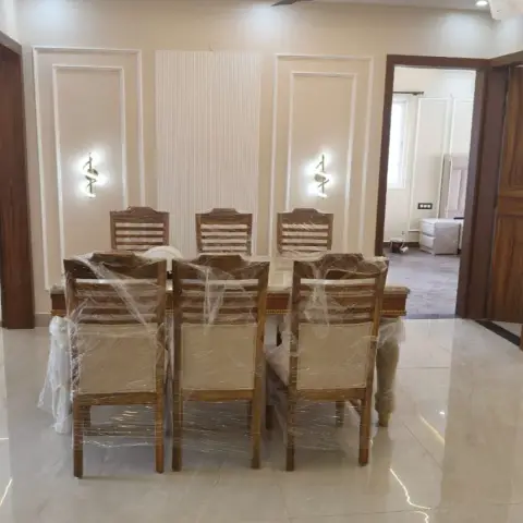 Six-seater teak dining set still in its delivery wrapping, set against a cream wall with two white moulding frames flanking a fluted panel and a pair of brass