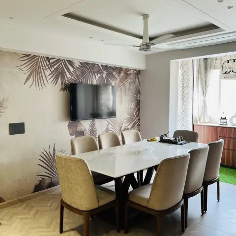 Six-seater white stone dining table on a dark X-frame base in front of a sepia tropical-leaf mural wall carrying a wall-mounted TV