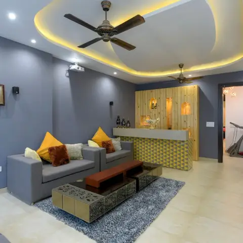Slate-blue lounge with a curved cove-lit ceiling loop carrying two antique-finish fans and a ceiling projector, grey sofas with mustard cushions on a shag rug