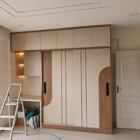 Sliding-door wardrobe with curved wood-grain inlay accents and a built-in study desk nook with lit overhead shelving, ladder and tools left from installation.