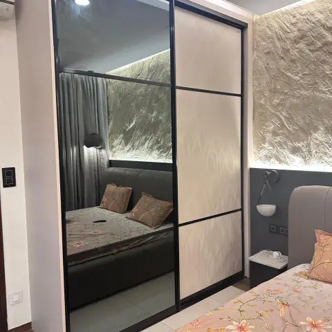 Sliding wardrobe in slim black profile frames — one bronze-tinted mirror panel reflecting the bed
