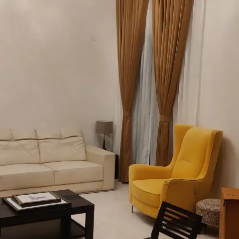 Sloping-ceiling living room with a dark exposed beam, cream leather three-seater, mustard velvet wingback and gold pinch-pleat curtains layered over white