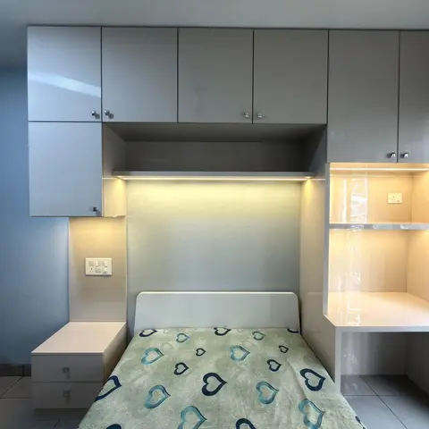 Small bedroom with overhead grey gloss storage cabinets, a bed with a heart-print bedspread, and a built-in study desk with warm LED task lighting.