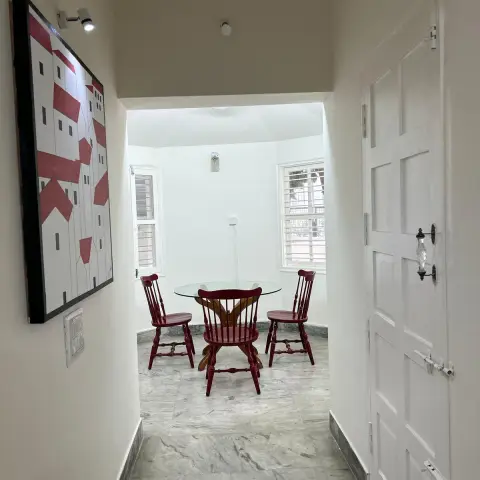 Small dining nook in a windowed bay at the end of a white passage - round glass-top table with three cherry-red spindle-back chairs on grey marble