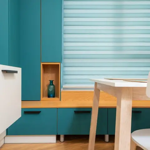 Small dining nook with a teal built-in bench and storage unit with a wood display niche, beside a wood table and sheer roller blinds.