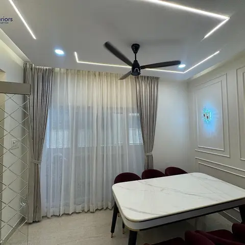 Small dining nook with a white marble-top table, maroon velvet chairs, sheer curtains and a panel-moulded accent wall with backlit wall art