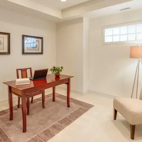 Small home study set up under a high strip window: a cherry-wood writing desk and side chair on a bordered beige rug
