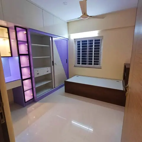 Small purple-and-cream room with a single box bed under the window, a sliding wardrobe with an open shelf bank