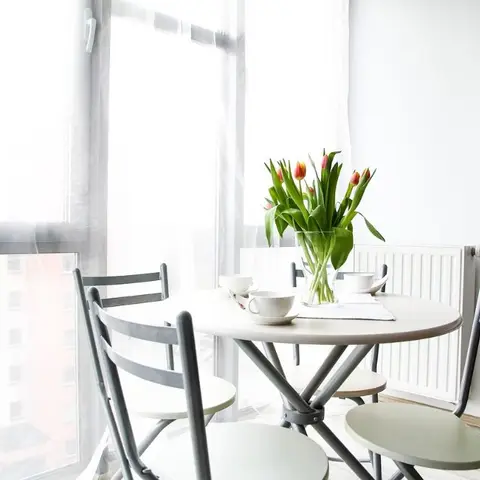 Small round dining table with folding metal-frame chairs by a sheer-curtained window, set with teacups and a vase of tulips